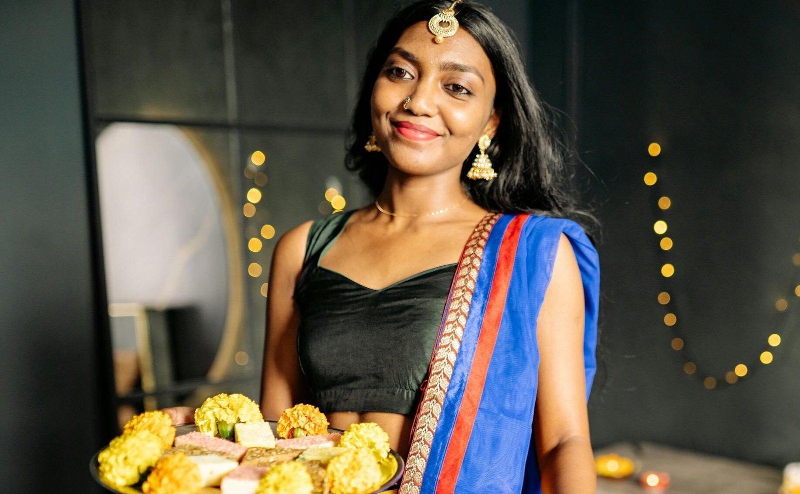 Smiling Indian woman in traditional attire holding a festive food platter indoors with decorative lights.